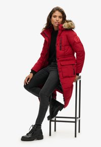 Abrigo largo acolchado rojo con capucha forrada de piel, múltiples bolsillos y detalles en cremallera negra. Jeans ajustados negros y botas negras resistentes con cordones.
