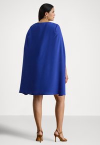 Kraljevsko modra, rahlo padajoča obleka s plaščem, s kvadratnim izrezom in ohlapnim krojem. Gladek tekstura, dolžina do kolen, združena z rjavimi sandali s tankimi trakovi.