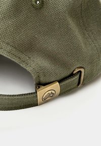 Gorra verde olivo hecha de tela texturizada con un broche metálico que presenta un logo. Correa ajustable y detalles de costura reforzada.