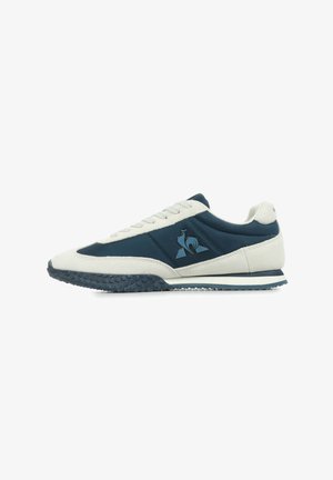 Zapatilla deportiva azul y crema con suela de goma texturizada, parte superior de ante y sintético, puntera redonda y detalle del logo en el lateral.