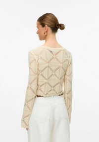 Beige gehaakte blouse met lange mouwen, geometrische patronen, open brei textuur, gekartelde zoom en aansluitend ontwerp, gecombineerd met witte broek.