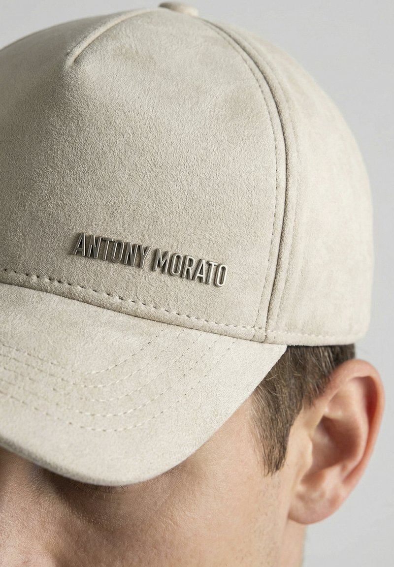 Lichtbeige suède baseballpet met metalen logo "ANTONY MORATO", gedragen op het hoofd van een man, waarbij zijn oor en gedeeltelijk gezicht zichtbaar zijn.