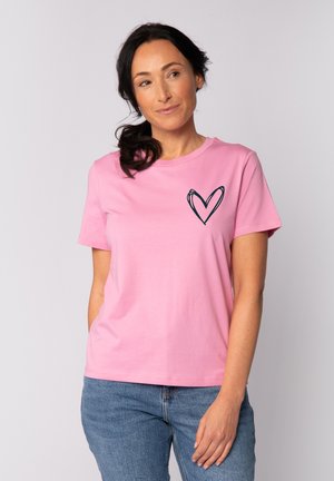 Frau mit dunklen Haaren im Pferdeschwanz, die ein rosa T-Shirt mit einem schwarzen Herzmuster und blaue Jeans trägt, steht vor einem einfarbigen Hintergrund.