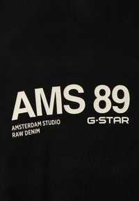 Schwarzer Stoff mit großem weißem Text: "AMS 89 AMSTERDAM STUDIO RAW DENIM G-STAR." Glatte Oberfläche mit matter Optik.