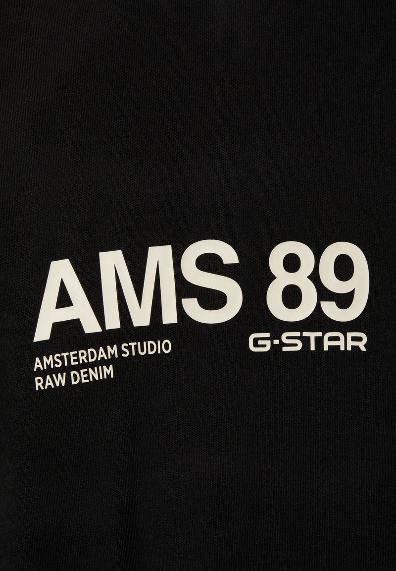Tessuto nero con grande testo bianco: "AMS 89 AMSTERDAM STUDIO RAW DENIM G-STAR." Superficie liscia con finitura opaca.