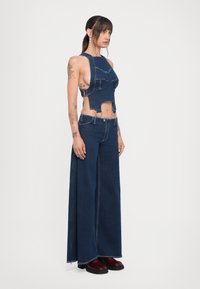 Denim odijelo s izrezanim halter crop topom s velikim džepovima i širokim hlačama, sve u tamnoplavoj boji. Teksturirana tkanina i jedinstven dizajn.