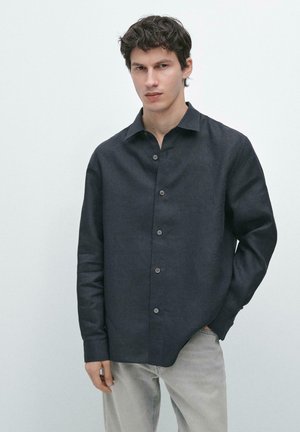 Junger Mann mit dunklem lockigem Haar, der ein dunkelgraues Hemd mit Knöpfen und hellgraue Hose trägt, vor einem schlichten weißen Hintergrund stehend.