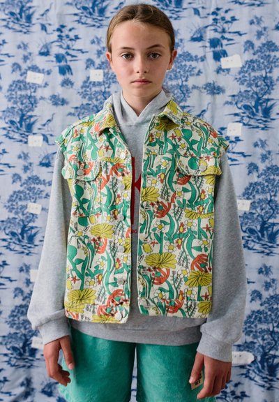 Młoda osoba ubrana w kolorową kamizelkę w kwiaty na szarą bluzę z kapturem i turkusowe spodnie, stojąca na tle niebiesko-białego wzoru.