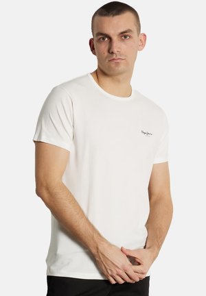 Giovane uomo che indossa una maglietta bianca a maniche corte con un piccolo logo nero "Pepe Jeans" sul petto a sinistra, in piedi davanti a uno sfondo neutro.