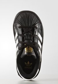 Svart Adidas-sneaker med vita ränder, guldlogotyp på tungan och innersulan, samt strukturerad skalmossdesign, visad från ovan.