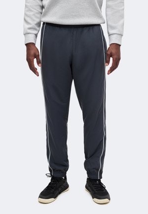 Mann trägt dunkelgraue Jogginghose mit weißen Seitenstreifen und schwarze Sneaker mit weißen Sohlen, steht vor einfachem Hintergrund.