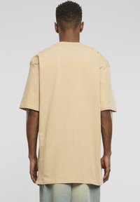 Beige oversized tričko s krátkým rukávem, které má kulatý výstřih a hladkou texturu. Zadní pohled ukazuje čisté linie a švy.
