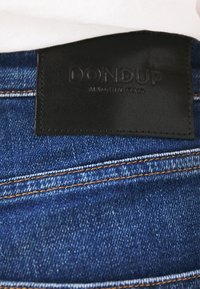 Jean en denim avec une étiquette en cuir noir embossée de "DONDUP" et "MADE IN ITALY". Caractérisé par des coutures orange et une surface de tissu texturée.