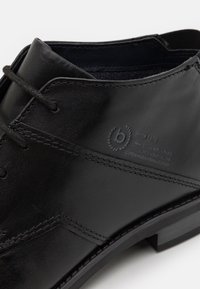 Chaussure noire en cuir avec une texture lisse, dotée d'un design à lacets, de coutures décoratives et d'un détail de logo près de la cheville.