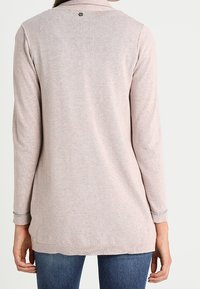 Žena nosící světle růžový kardigan s dlouhým rukávem a límečkem, viděná ze zadu, v kombinaci s modrými džíny.