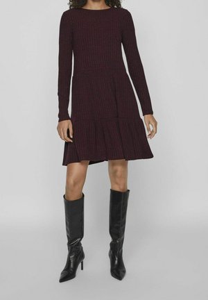 Femme portant une robe côtelée à manches longues de couleur violet foncé avec une jupe à étages et des bottes noires à talons montant jusqu'aux genoux, debout sur un fond gris.