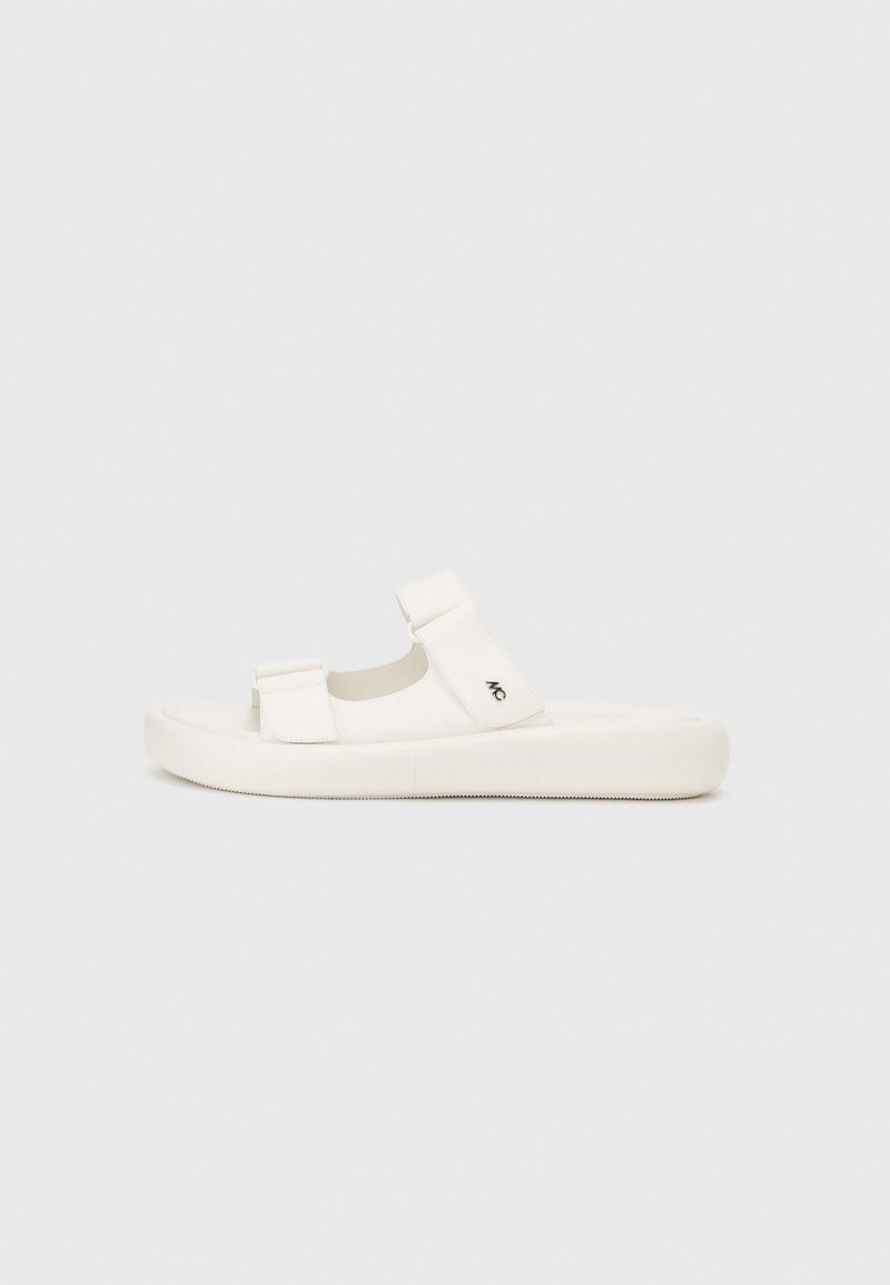 Witte sandalen met twee verstelbare banden, glad materiaal, platte zool, ronde neus en een logo-detail aan de zijkant. Minimalistisch ontwerp.