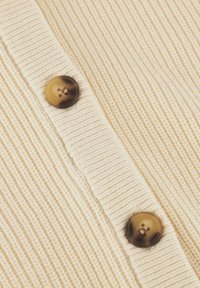 Beige geribbelde cardigan met twee donkere, ronde knopen langs de opening. Materiaal heeft een zachte, textuurafwerking.