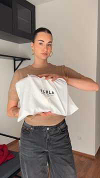 Νεαρή γυναίκα με μαλλιά μαζεμένα και χρυσά σκουλαρίκια σε στιλ κρίκου κρατάει μια λευκή θήκη Furla σε έναν σύγχρονο εσωτερικό χώρο.