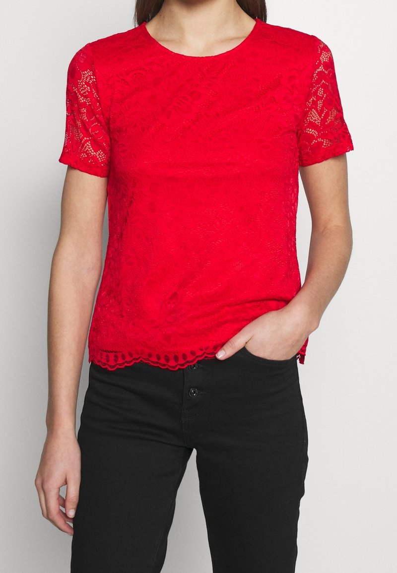 Femme portant un haut en dentelle rouge vif à manches courtes et un pantalon noir, debout avec une main dans la poche devant un fond uni.