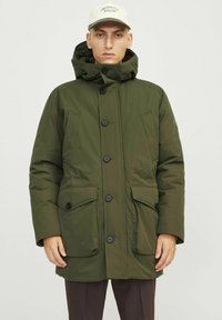 Parka acolchada de color verde oliva con capucha, con botones negros, dos bolsillos frontales y una textura suave. Acentuada con una gorra de color claro.