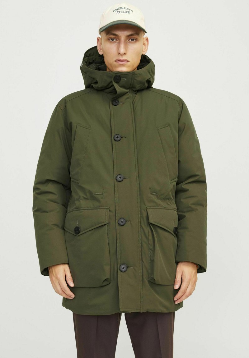Parka acolchada de color verde oliva con capucha, con botones negros, dos bolsillos frontales y una textura suave. Acentuada con una gorra de color claro.