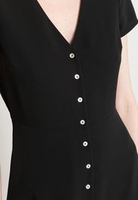 Vestido preto com decote em V, mangas curtas e uma linha de seis botões pérola na parte da frente. Tecido liso e leve.