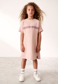 Šviesiai rožinė medvilninė suknelė su trumpomis rankovėmis, ant krūtinės išsiuvinėta violetinė juosta. Derinama su baltais sportbačiais ir kojinėmis.