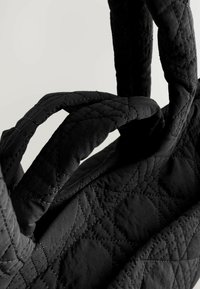 Schwarze gesteppte Handtasche mit zwei stabilen Griffen, die den strukturierten Stoff und die Nähdetails vor einem neutralen Hintergrund betont.