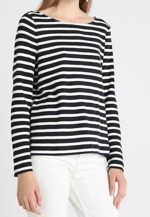 Frau trägt ein langärmliges, schwarz-weiß gestreiftes Oberteil mit waagerechten Streifen und weiße Hose, steht vor einem schlichten, hellen Hintergrund.