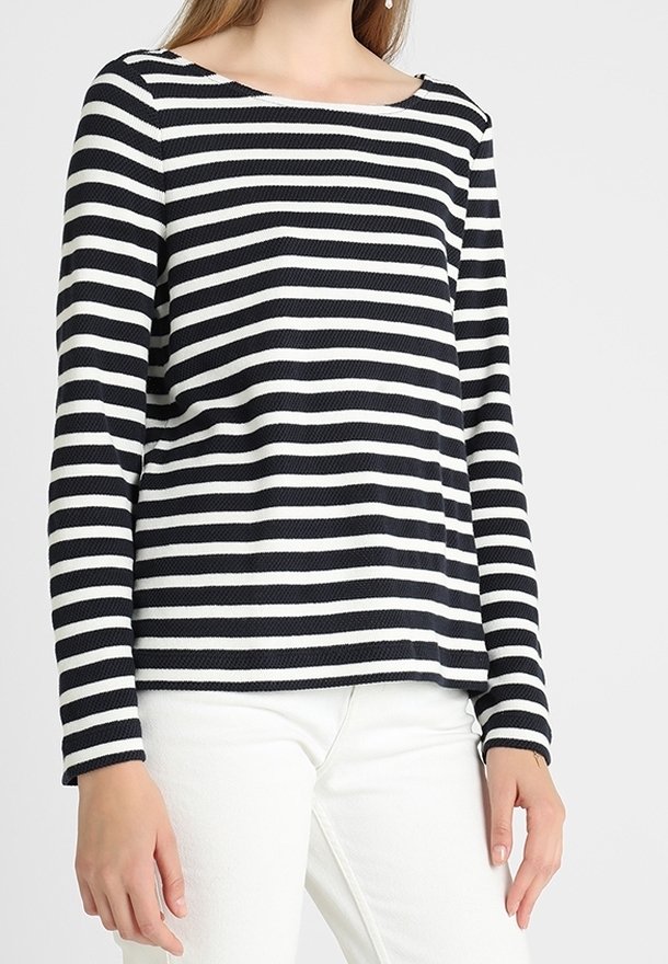 Femme portant un haut à rayures horizontales noires et blanches à manches longues avec un pantalon blanc, debout devant un fond clair uni.