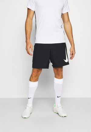 Mężczyzna - sportowiec w białej koszulce sportowej, czarnych spodenkach z logo, białych podkolanówkach i białych korkach piłkarskich z zielonymi akcentami.