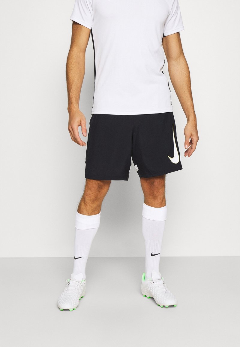 Athlète masculin portant un maillot de sport blanc, un short noir avec un logo, des chaussettes blanches montantes et des chaussures de football blanches avec des accents verts.