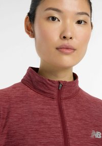 Maroon zip-up shirt with a high collar, featuring a textured fabric and a small silver logo on the left side. Closely fitted design.