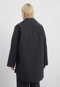 Cămașă lungă neagră cu guler, realizată dintr-un material texturat. Are un croi drept, slituri laterale și cusături cu umeri lăsați pentru confort.