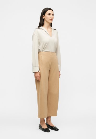 Γυναίκα όρθια, κοιτάζοντας μπροστά, φοράει κρεμ τοπ με V-λαιμόκοψη και μακριά μανίκια, μπεζ παντελόνι με φαρδιά μπατζάκια και ψηλή μέση και μαύρα φλατ, σε λευκό φόντο.