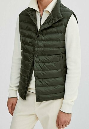 Homme portant un gilet matelassé sans manches vert foncé sur une chemise blanche à manches longues avec fermeture éclair et un pantalon crème, debout devant un fond uni.