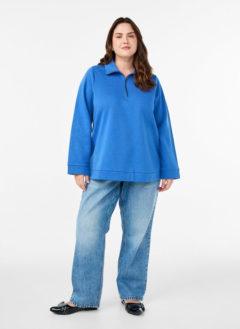 Vrouw staat in een blauwe lange mouwen trui met rits, lichtblauwe losse spijkerbroek en zwarte platte schoenen met gespsluiting.