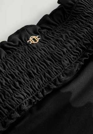 Détail en tissu noir présentant une texture froncée avec des bords volants, orné d'un accent circulaire doré en haut.