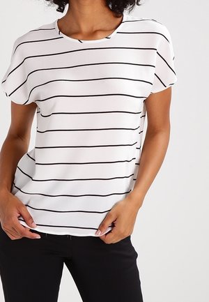 Femme portant un haut blanc à manches courtes avec des rayures horizontales noires, associée à un pantalon noir, debout devant un fond uni.