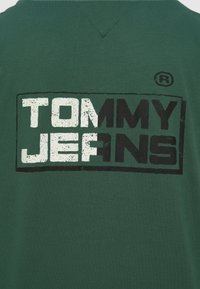 Πράσινο βαμβακερό μπλουζάκι με ορθογώνιο ασπρόμαυρο λογότυπο "TOMMY JEANS" στην πλάτη, με υφή και σχέδιο σε σχήμα V στη λαιμόκοψη.