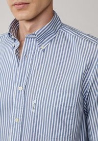 Camisa de botones a rayas azules y blancas con un cuello clásico, bolsillo en el pecho y botones blancos. Fabricada con tela ligera de textura suave.