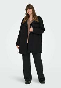 Černý oversized kabát s dvojitými knoflíky, širokými klopami a měkkou tkaninou. Stylizováno s béžovým topem a volnými černými kalhotami.