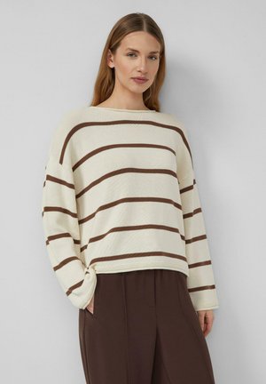 Donna con capelli lunghi che indossa un maglione crema con strisce orizzontali marroni e pantaloni a gamba larga di colore marrone scuro, in piedi davanti a uno sfondo semplice.