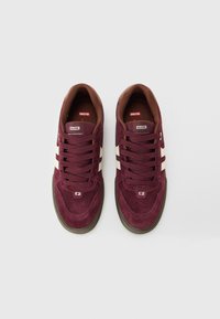 Ein Paar burgunderfarbene Wildleder-Sneaker mit weißen Seitensch stripes und braunen Sohlen, von oben auf einem schlichten weißen Hintergrund betrachtet.