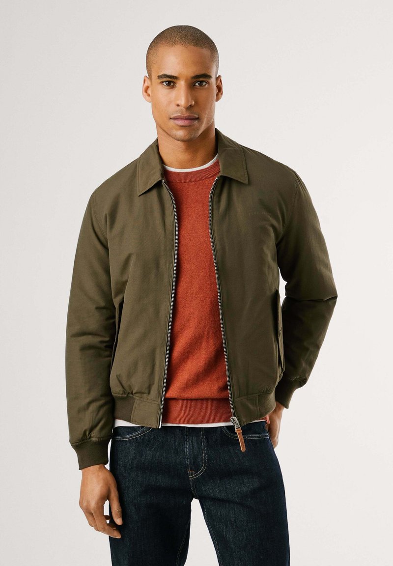 Chaqueta bomber verde oliva con cierre de cremallera y cuello, superpuesta sobre un suéter de color óxido. Combinada con jeans oscuros, textura suave en toda la prenda.