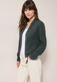 Dunkelgrüne Bomberjacke aus strukturiertem Stoff, Reißverschluss und zwei Taschen. Kombiniert mit einem weißen Oberteil und hellfarbigen Hosen.