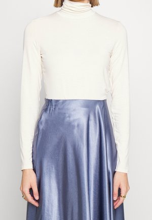 Vrouw draagt een witte, lange mouw coltrui, ingestopt in een hooggetailleerde, glanzende lavendelkleurige satijnen rok, handen langs het lichaam.