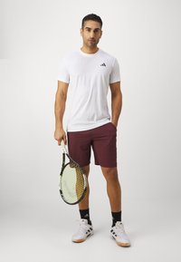 T-shirt de sport blanc à finition texturée, shorts bordeaux et raquette de tennis, avec des accents noirs et des chaussures de sport blanches.
