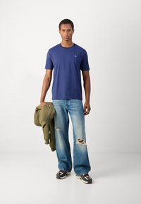 Mørkeblå bomulds t-shirt med korte ærmer og et lille logo, parret med lyseblå slidte jeans og en olivengrøn jakke.